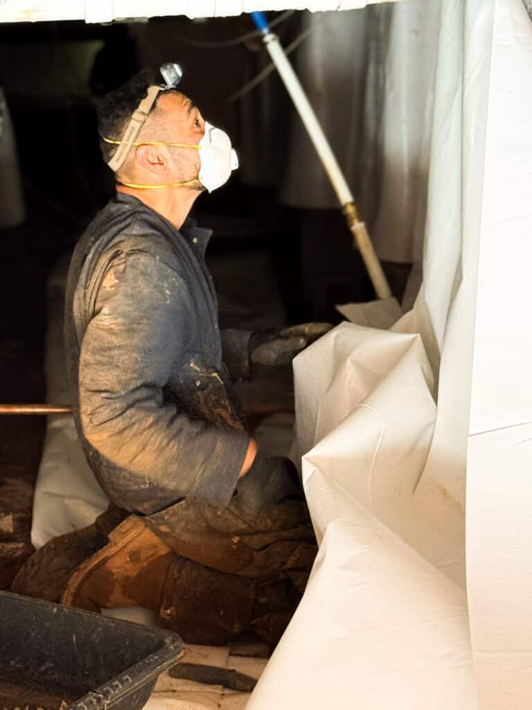 A crew member is installing vinyl sheeting on the walls within the crawl space.